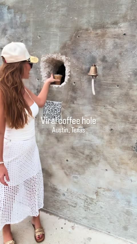 Woman reaching into a hole in a concrete wall at A Hole Coffee Austin to take her drink, bell beside it.